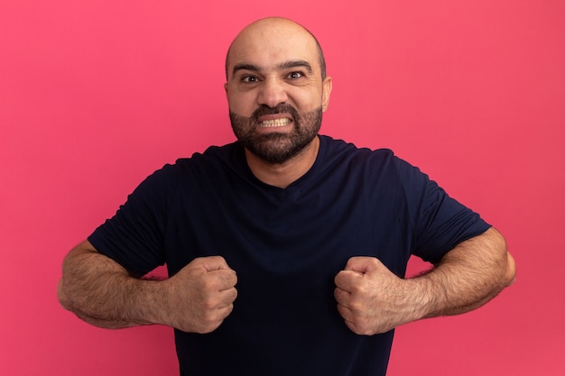 Homem barbudo em uma camiseta azul marinho com punhos cerrados, feliz e animado em pé sobre uma parede rosa