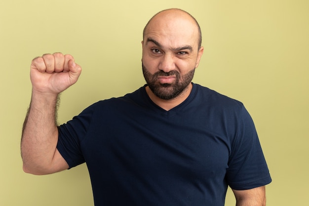 Homem barbudo em uma camiseta preta com rosto carrancudo levantando o punho como um vencedor em pé sobre uma parede verde
