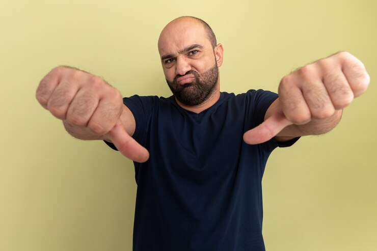 Foto grátis homem barbudo em uma camiseta preta insatisfeito mostrando os polegares para baixo em pé sobre uma parede verde