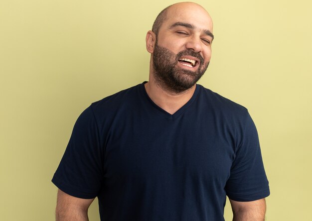 Homem barbudo em uma camiseta preta sorrindo com uma carinha feliz em pé sobre a parede verde