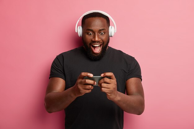 Homem barbudo empolgado segura o smartphone moderno horizontalmente, joga jogo online, usa o conjunto de cabeça, preso na tela do gadget, vestido com uma camiseta preta, se diverte sozinho
