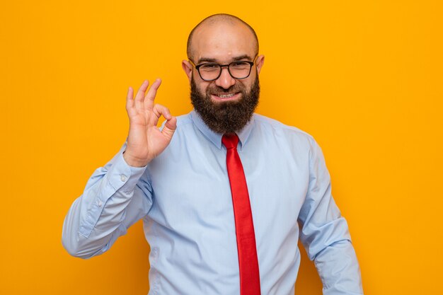 Homem barbudo feliz com gravata vermelha e camisa azul, usando óculos, sorrindo e mostrando sinal de ok