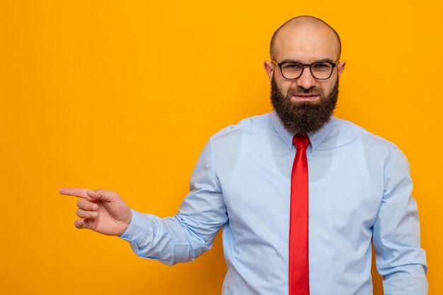 Homem barbudo feliz com gravata vermelha e camisa usando óculos, olhando para a câmera apontando com o dedo indicador para o lado e sorrindo confiante em pé sobre um fundo laranja