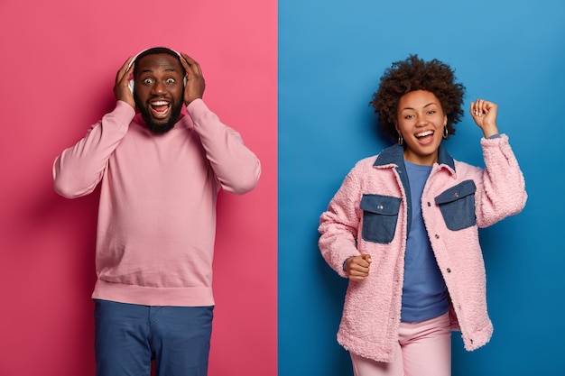 Homem barbudo feliz usa fones de ouvido estéreo, canta e atinge a nota mais alta, mulher alegre e despreocupada dança perto, mantém os braços erguidos, isolado sobre uma parede rosa e rosa.