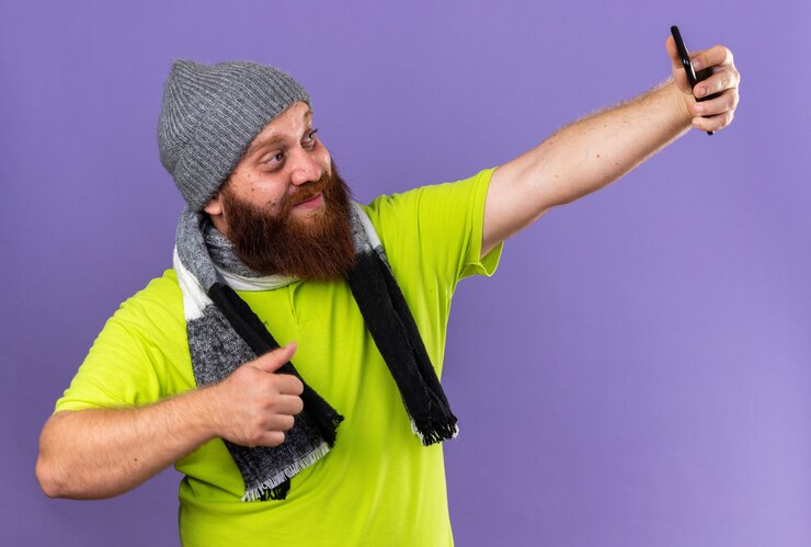 Foto grátis homem barbudo insalubre com chapéu e cachecol quente no pescoço sorrindo e se sentindo melhor fazendo selfie usando smartphone