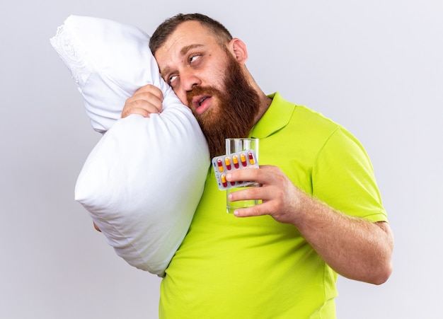 Foto grátis homem barbudo insalubre em uma camisa pólo amarela com travesseiro segurando um copo d'água e comprimidos, sentindo-se doente, sofrendo de gripe, em pé sobre uma parede branca