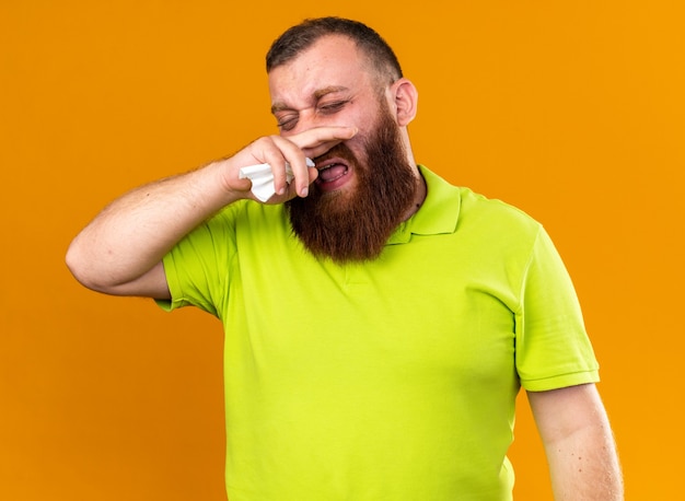 Homem barbudo insalubre em uma camisa pólo amarela se sentindo terrível, sofrendo de frio, enxugando o nariz escorrendo em pé sobre uma parede laranja