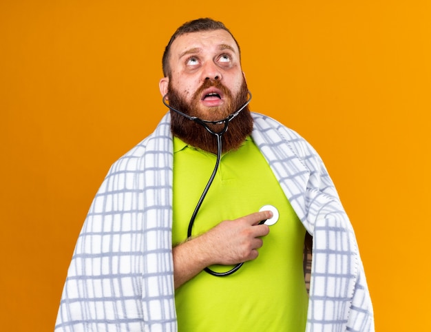 Foto grátis homem barbudo insalubre envolto em cobertor, sentindo-se mal, sofrendo de frio, ouvindo os batimentos cardíacos usando um estetoscópio