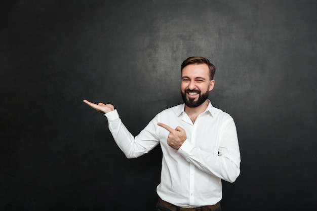 Homem barbudo otimista apontando o dedo indicador, mantendo a coisa na palma da mão, demonstrando ou anunciando sobre o espaço da cópia cinza escuro