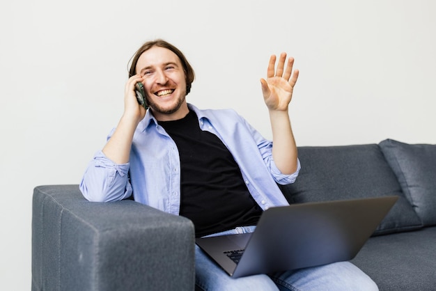Homem barbudo positivo com laptop falando no smartphone sentado no sofá na sala de estar