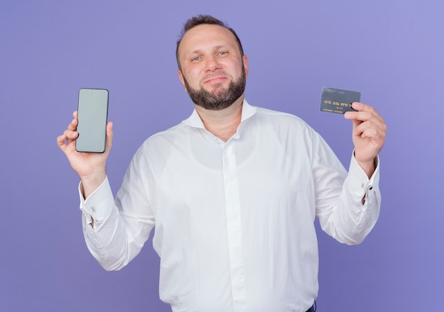 Homem barbudo satisfeito vestindo camisa branca mostrando smartphone e cartão de crédito sorrindo em pé sobre a parede azul