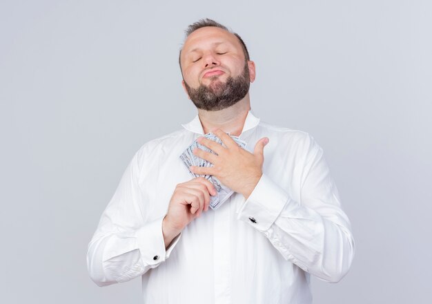 Homem barbudo satisfeito, vestindo uma camisa branca e segurando dinheiro, parecendo confiante em pé sobre uma parede branca