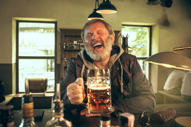 Homem barbudo sênior bebendo álcool em um bar e assistindo a um programa de esporte na TV. Aproveitando minha cerveja favorita. Homem com uma caneca de cerveja, sentado à mesa. Fã de futebol ou esporte. Conceito de emoções humanas