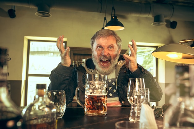 Homem barbudo sênior bebendo álcool em um bar e assistindo a um programa de esporte na tv.