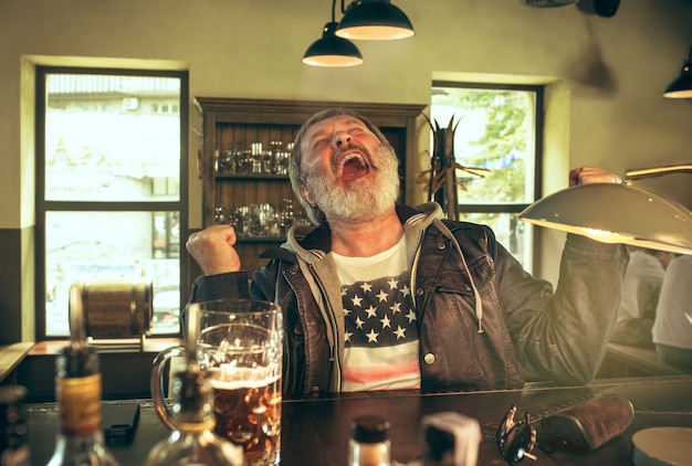 Homem barbudo sênior bebendo álcool em um bar e assistindo a um programa de esporte na TV.