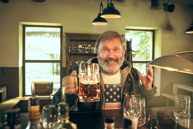 Homem barbudo sênior bebendo álcool em um bar e assistindo a um programa de esporte na tv.