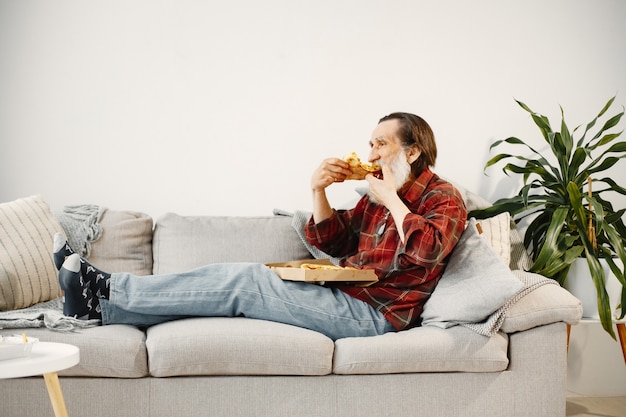 Homem barbudo sênior deitado no sofá e comendo pizza. Comida rápida.