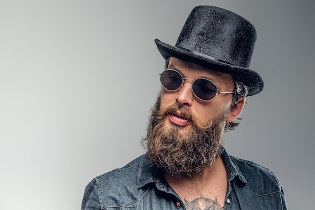 Homem barbudo sério de chapéu e óculos de sol está posando no estúdio fotográfico.
