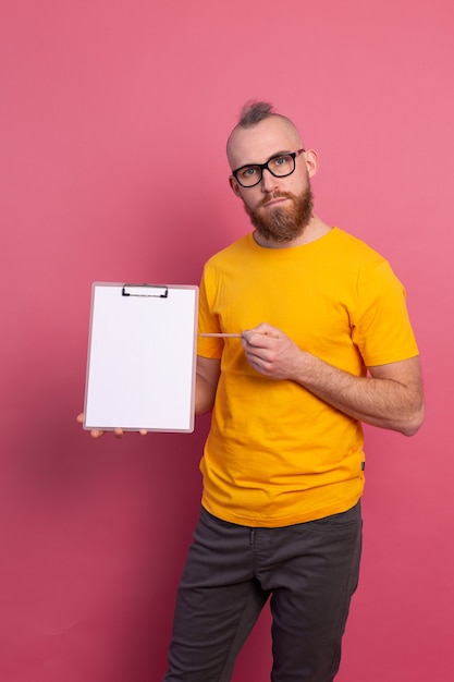 Homem barbudo sorridente usando óculos e roupas casuais segurando uma prancheta com um papel em branco apontando