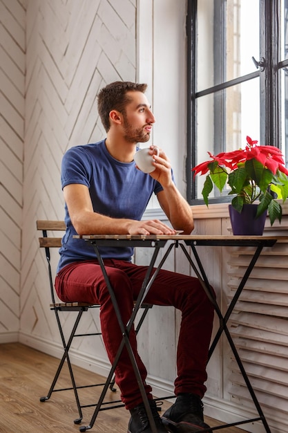 Homem barbudo tomando café na mesa perto da janela.