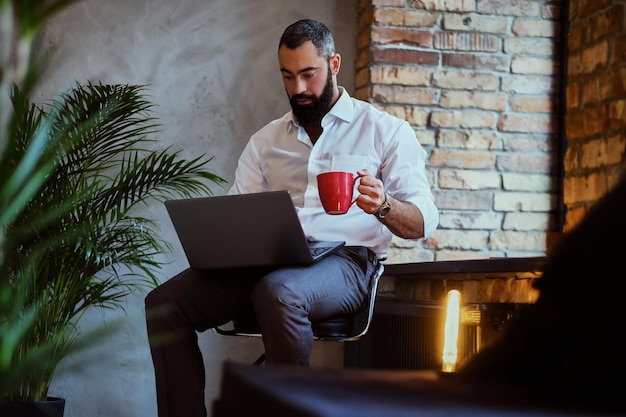 Homem barbudo urbano bebe café e usando um laptop em uma sala com interior loft.