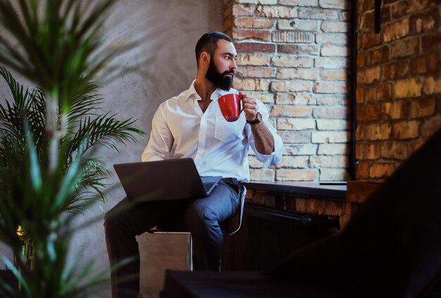 Homem barbudo urbano bebe café e usando um laptop em uma sala com interior loft.