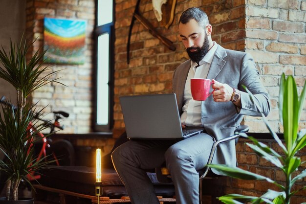 Homem barbudo urbano bebe café e usando um laptop em uma sala com interior loft.