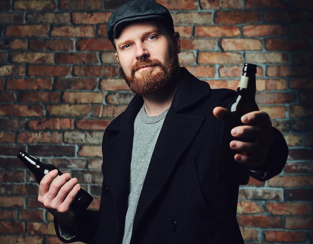 Foto grátis homem barbudo vestido com um boné de tweed preto e jaqueta de lã segura garrafa de cerveja sobre a parede de um tijolo.