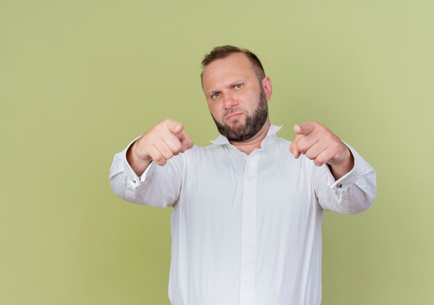 Homem barbudo vestindo camisa branca apontando com os dedos indicadores e parecendo descontente em pé perto da parede de luz