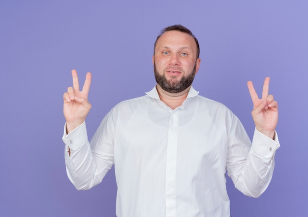 Foto grátis homem barbudo vestindo camisa branca e sorrindo mostrando o sinal v em pé sobre a parede azul