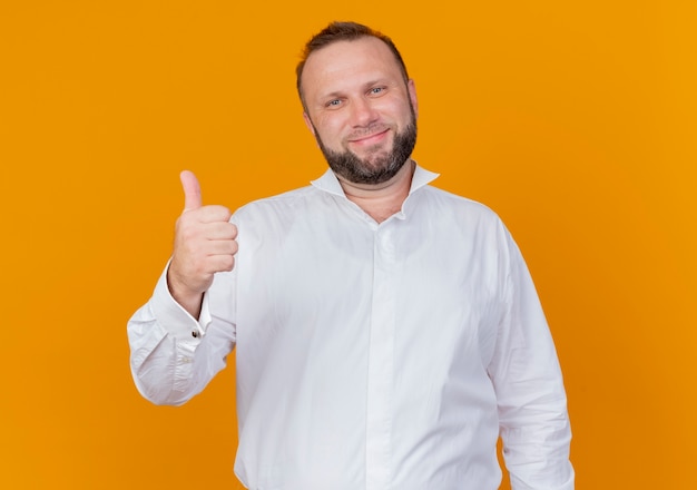 Homem barbudo vestindo camisa branca e sorrindo mostrando os polegares em pé na parede laranja