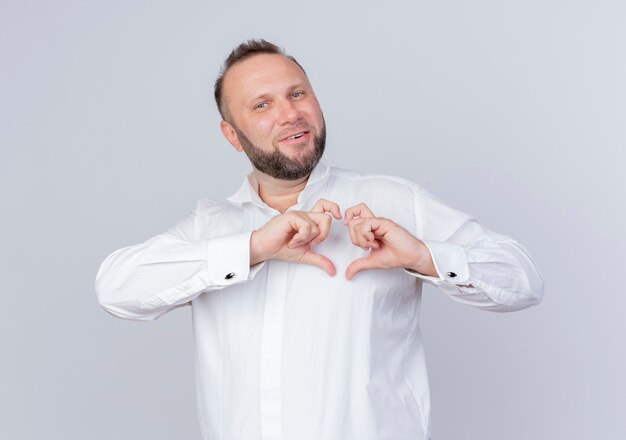 Homem barbudo vestindo camisa branca fazendo um gesto de coração com os dedos sorrindo em pé sobre uma parede branca