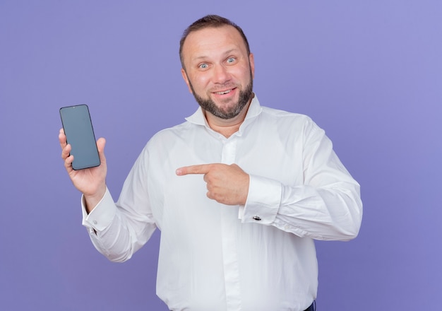 Homem barbudo vestindo camisa branca mostrando a ponta do smartphone com o dedo indicador sorrindo em pé sobre a parede azul