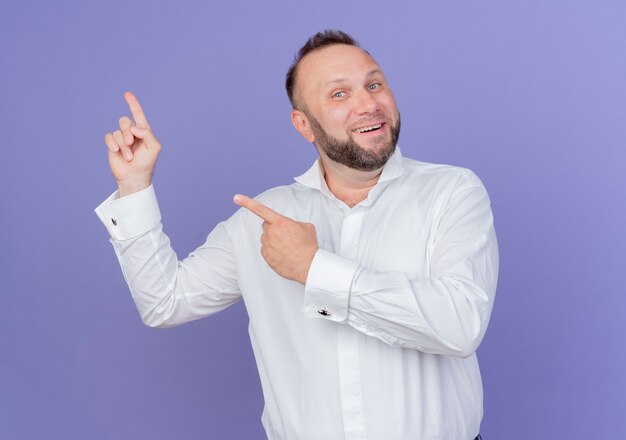 Homem barbudo vestindo camisa branca sorrindo com uma cara feliz apontando com os dedos para o lado em pé sobre a parede azul