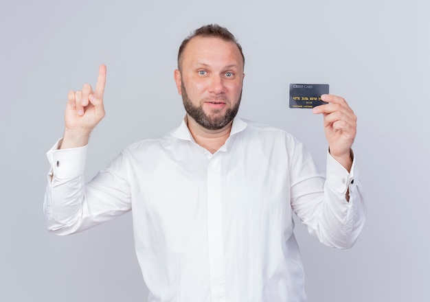Homem barbudo, vestindo uma camisa branca, mostrando o cartão de crédito, mostrando o dedo indicador e sorrindo com uma cara feliz, tendo uma nova ideia em pé sobre uma parede branca