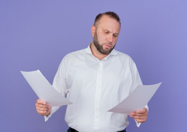 Homem barbudo vestindo uma camisa branca segurando folhas de papel em branco, olhando para elas com uma cara séria em pé sobre a parede azul