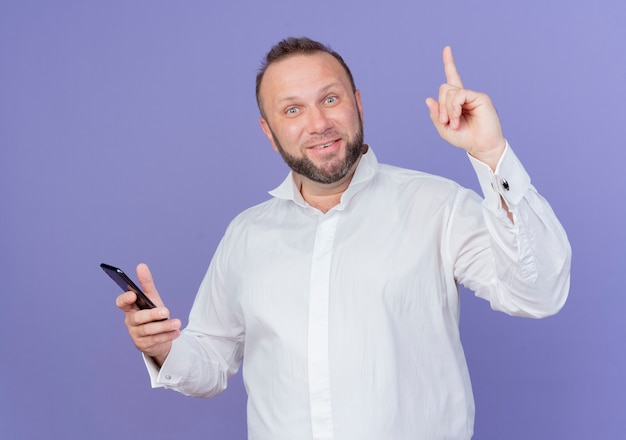 Homem barbudo, vestindo uma camisa branca, segurando um smartphone, parecendo feliz e surpreso, mostrando o dedo indicador tendo uma nova ideia em pé sobre a parede azul