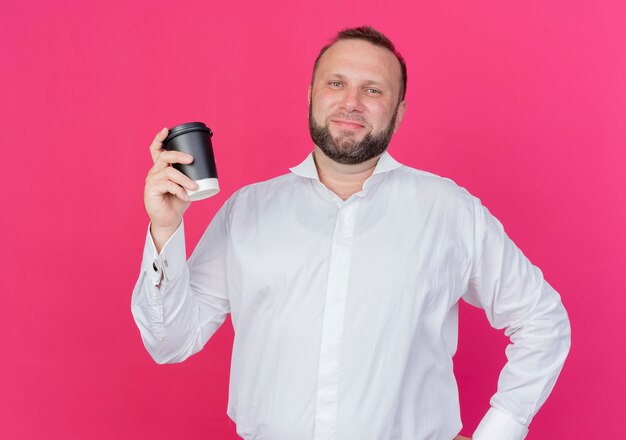 Homem barbudo, vestindo uma camisa branca, segurando uma xícara de café, parecendo amigável em pé sobre uma parede rosa