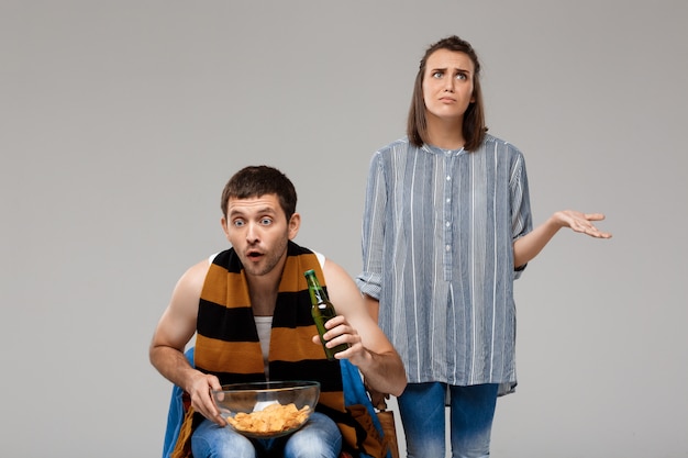 Homem bebendo cerveja, assistindo futebol e chateado mulher em pé atrás