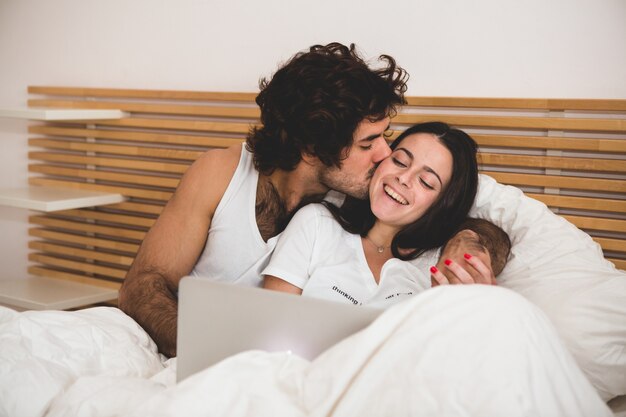 Homem beijando sua namorada enquanto eles olhar para um laptop na cama