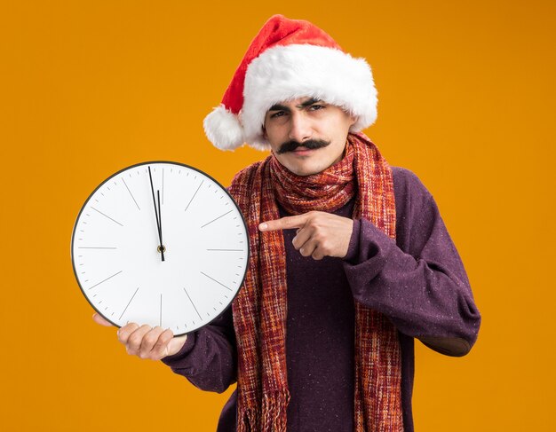 Foto grátis homem bigodudo usando chapéu de papai noel de natal com lenço quente em volta do pescoço segurando um relógio apontando com o dedo indicador para ele, parecendo confuso e descontente em pé sobre uma parede laranja