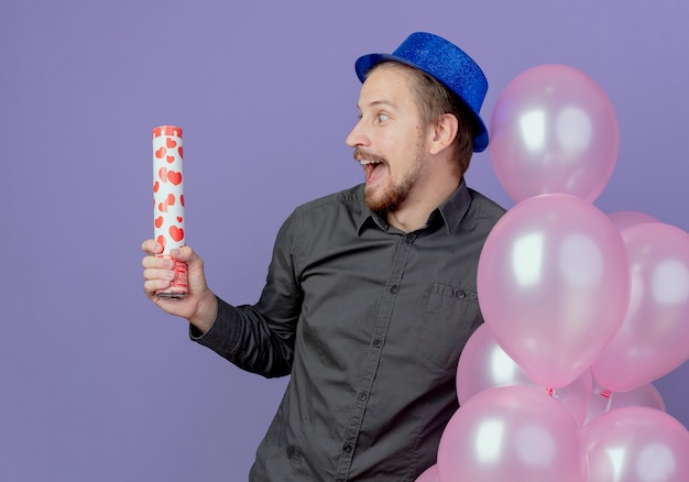 Foto grátis homem bonito animado com chapéu azul em pé com balões de hélio segurando e olhando para um canhão de confete isolado na parede roxa