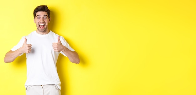 Homem bonito animado mostrando os polegares para cima, aprovando e dizendo que sim, de pé sobre um fundo amarelo