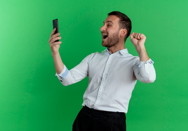 Homem bonito animado segurando e olhando para o telefone levanta o punho isolado na parede verde