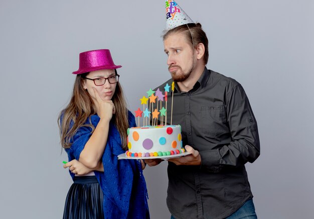 Homem bonito ansioso com um boné de aniversário segurando um bolo e olhando para uma jovem irritada de óculos com chapéu rosa segura o apito e coloca a mão no queixo