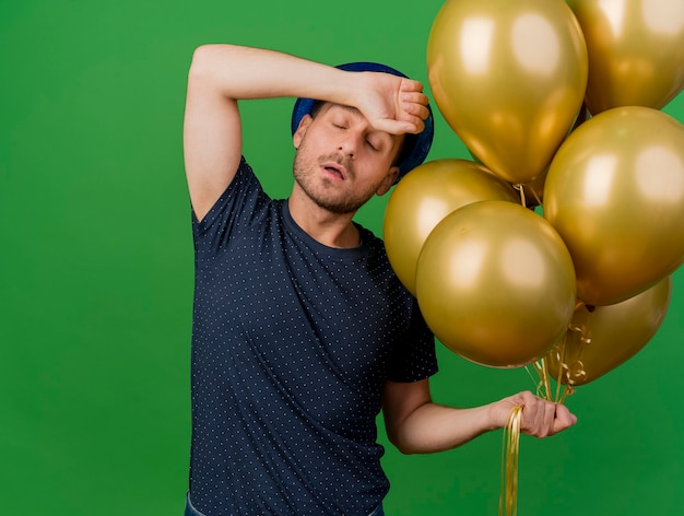 Homem bonito caucasiano irritado com chapéu de festa azul segura balões de hélio e coloca a mão na testa isolada em um fundo verde com espaço de cópia