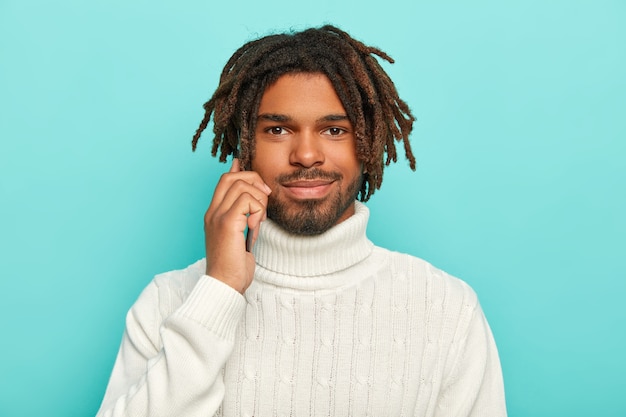 Homem bonito com a barba por fazer segura um celular moderno próximo ao ouvido, conversa ao telefone, usa um suéter branco quente, tem barba e dreadlocks