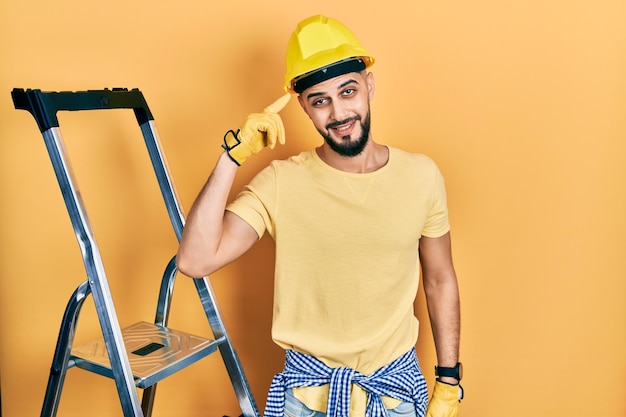 Foto grátis homem bonito com barba por escadas de construção usando capacete sorrindo apontando para a cabeça com um dedo ótima ideia ou boa memória