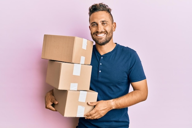 Homem bonito com barba segurando pacotes de entrega olhando positivo e feliz em pé e sorrindo com um sorriso confiante mostrando os dentes