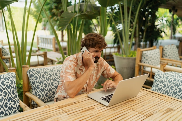 Homem bonito com barba usando lap top e telefone mobyle sentado no café ao ar livre com interior tropical